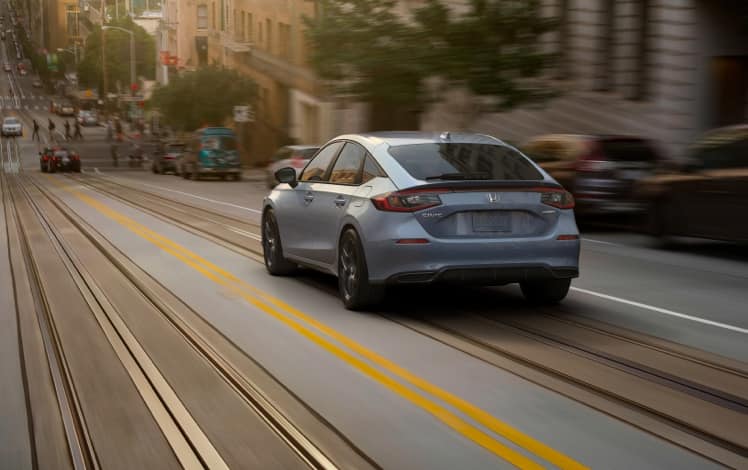Vista trasera desde el lado del conductor del Honda Civic Hatchback Sport 2023 en Sonic Gray Pearl conduciendo por una calle urbana.