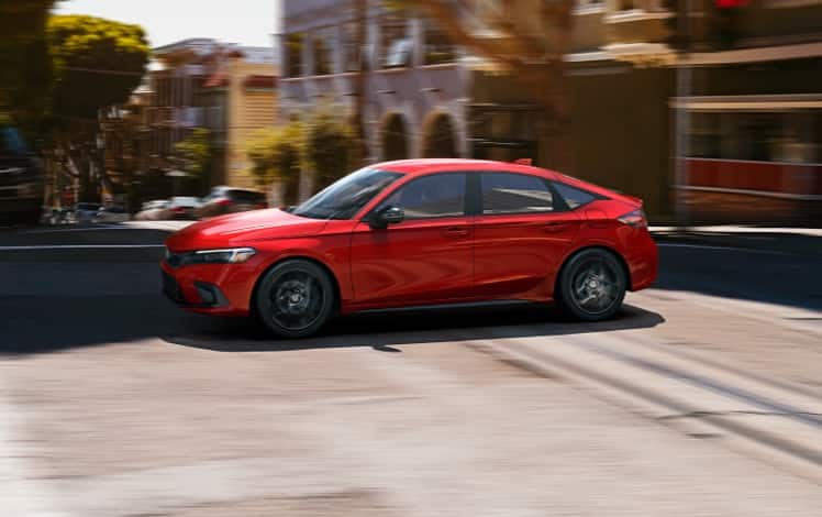 Vista de perfil desde el lado del conductor del Honda Civic Hatchback Sport 2023 en Rallye Red conduciendo por una calle de ciudad.