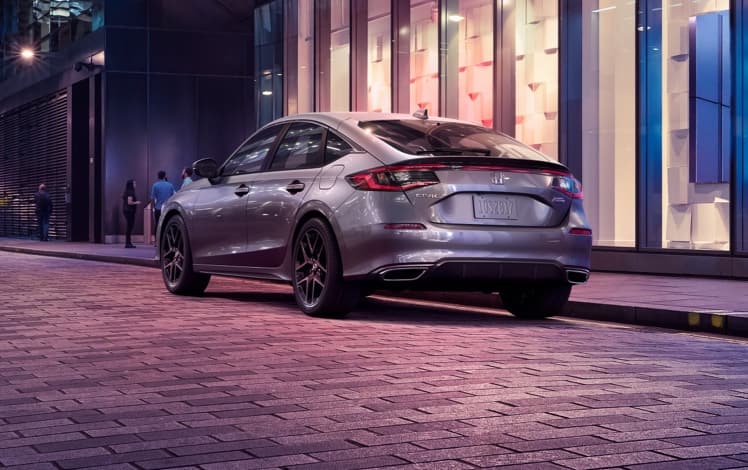 Vista trasera desde el lado del conductor del Honda Civic Sport Touring Hatchback 2023 en Lunar Silver Metallic estacionado afuera de una tienda.